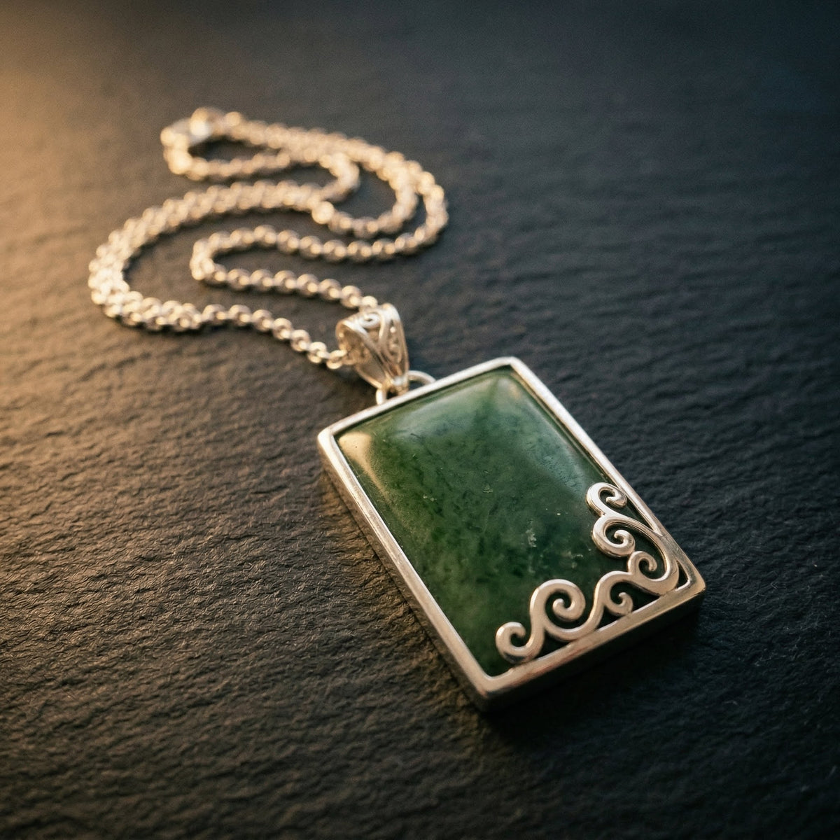 Rectangular pounamu pendant in silver setting with carved koru detailing along lower edge, resting on dark timber grain with warm side light catching the silver chain. A considered NZ-made gift for someone meaningful — pounamu jewellery gift from Aotearoa.