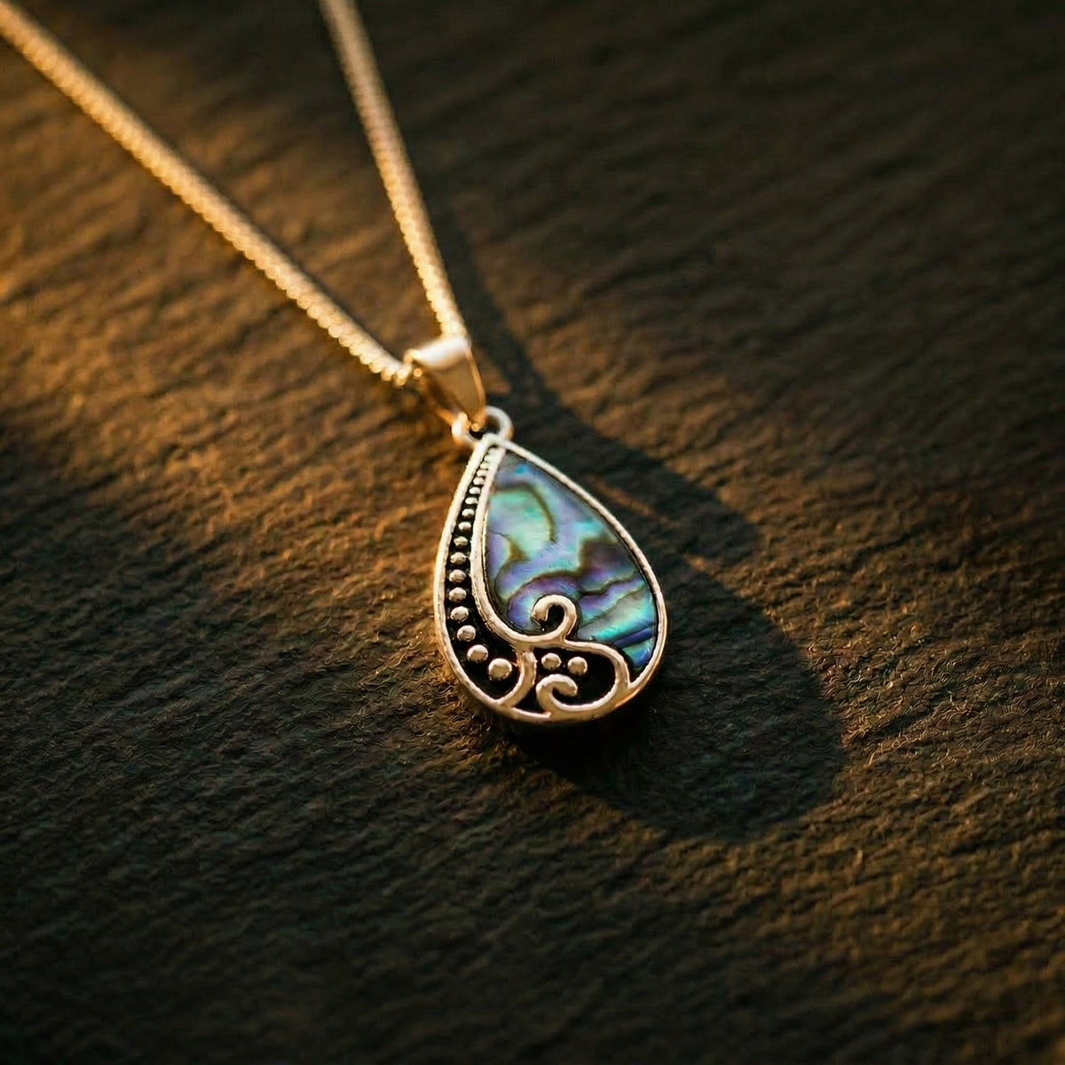 Silver teardrop pendant with paua shell inlay and koru detailing, on fine chain resting across dark timber in warm afternoon light. A considered NZ-made gift for Mum or someone meaningful — arrives ready to gift in an EMBER box.