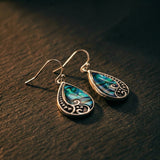 Silver teardrop earrings with paua shell inlay, blue-green iridescence catching warm light on dark timber.