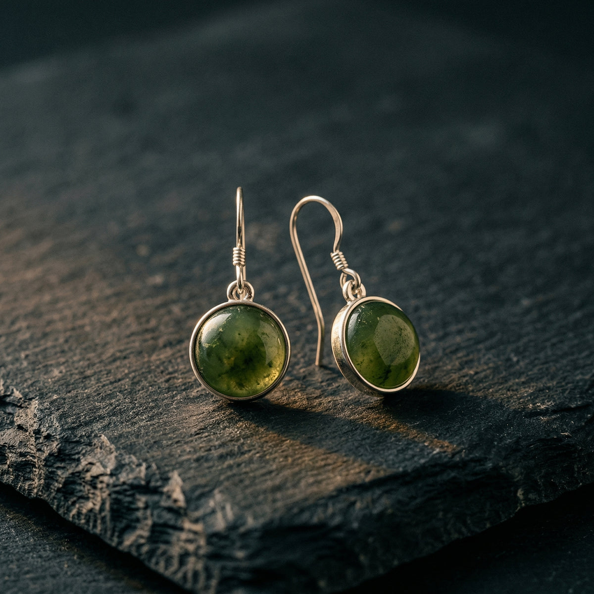 Silver drop earrings with round pounamu stones, forest green with amber inclusions, on dark slate with soft fern shadow. A considered NZ-made gift for someone meaningful — pounamu jewellery for Mum, a milestone birthday or thank you gift NZ.