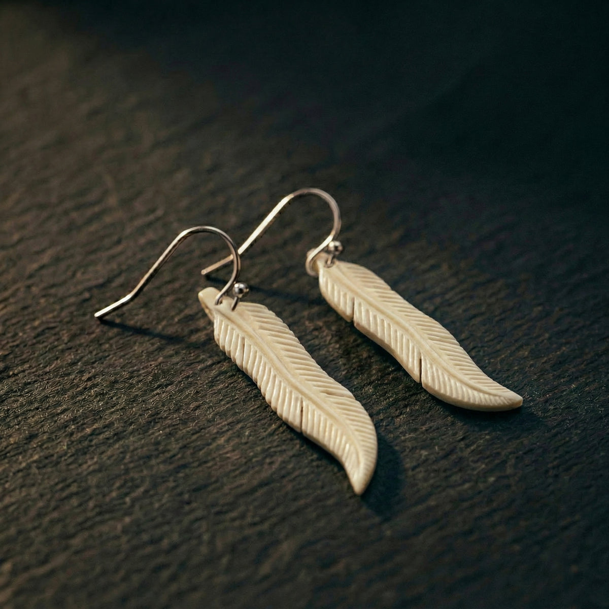 Carved bone feather earrings on sterling silver hooks, resting on dark timber grain with soft light catching the detail. A considered NZ-made gift for someone who has everything — worn, meaningful jewellery that carries quiet presence.