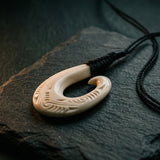 Carved bone hei matau pendant on black woven cord, resting against dark slate with soft natural light and traditional Māori koru patterns. A considered NZ-made gift for someone meaningful — unique jewellery that carries cultural weight.