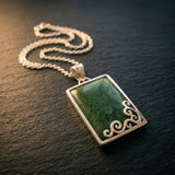 Rectangular pounamu pendant in silver setting with carved koru detailing along lower edge, resting on dark timber grain with warm side light catching the silver chain. A considered NZ-made gift for someone meaningful — pounamu jewellery gift from Aotearoa.